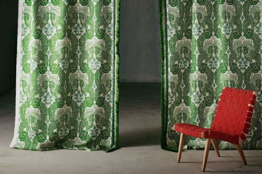 Drapery curtains in Belle-de-Nuit printed fabric in green and white linen with blue leaves, featuring a floral leaf design, by Raoul Dufy for Christopher Farr Cloth. Printed onto white linen in the UK. With green trims, bigtop tape, and ombre fringe edge, red chair in front, set in a concrete room. Photography by Christopher Horwood.