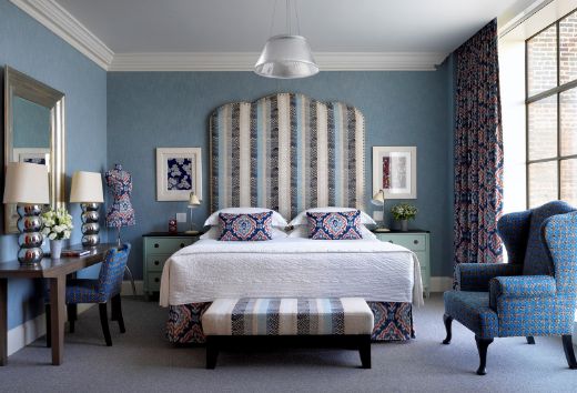 Eclectic hotel room at Crosby Street, Firmdale Hotel, featuring a Bookend Embroidered Fabric navy headboard with striped embroidery and studded edges. The matching footstool and red and blue accents in accessories and ikat-patterned curtains complete the design, photographed by Simon Brown.