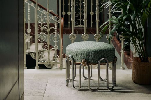 Decorative ornate footstool upholstered in olive Chiselled Circle fabric by And Objects for Christopher Farr Cloth, placed in an ornate hallway with metal decorative banisters.