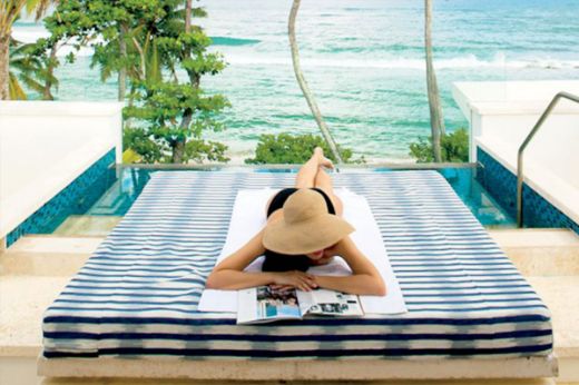 Woman reading a magazine on a poolside bed upholstered in Capri Outdoor Woven Fabric Azzurro, with sea view in the background