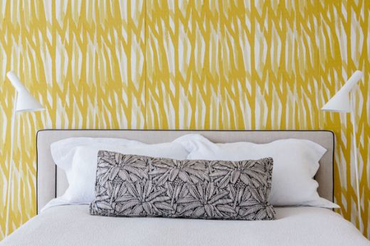 Bedroom interior featuring Christopher Farr Breakwater wallpaper in Mimosa yellow behind a bed dressed in crisp white linen, accented with a black and white palm tree bolster cushion. A fresh, modern look blending bold pattern with clean, tropical-inspired details.