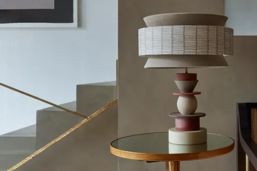 Vorster & Braye Totum ceramic lamp base in natural clay on a glass side table with a gold edge, featured in Christopher Farr Cloth's showroom designed by Hubert Zandberg. The lamp is paired with a bespoke neutral decorative-shaped lampshade, enhancing the modern, sleek interior.