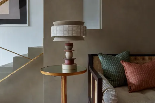 Vorster & Braye Totum ceramic lamp base in natural clay on a glass side table with a gold edge, featured in Christopher Farr Cloth's showroom designed by Hubert Zandberg. The lamp is paired with a bespoke neutral decorative-shaped lampshade, enhancing the modern, sleek interior. next to a cane sofa and striped algonquin cushions in green and orange for a pop of colour.